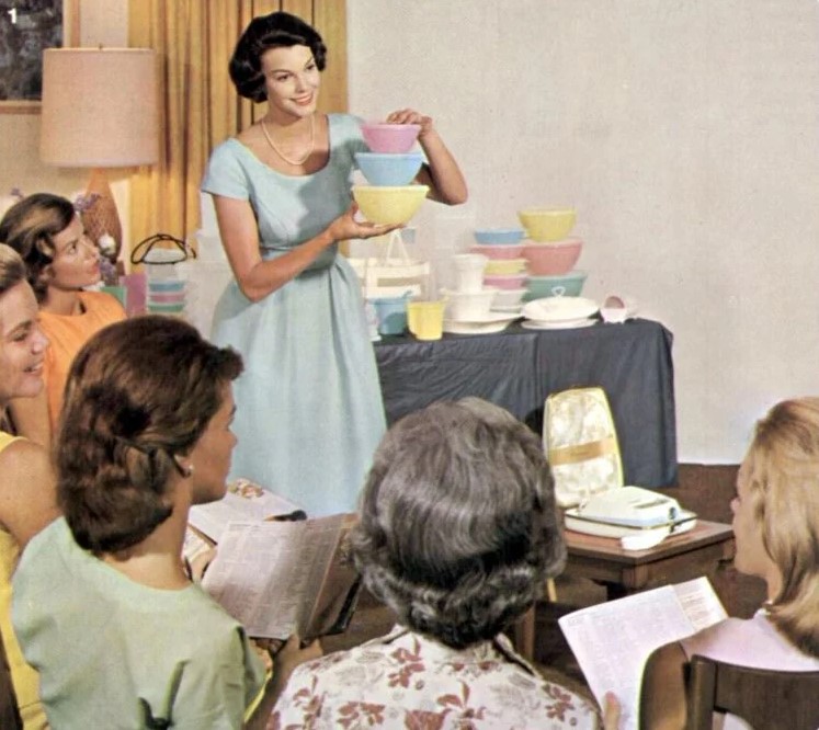 A woman in a blue dress shows tupperware containers to a group of women.