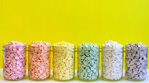 Smarties candies in six jars from left to right, pink, orange, yellow, green, white, and purple.