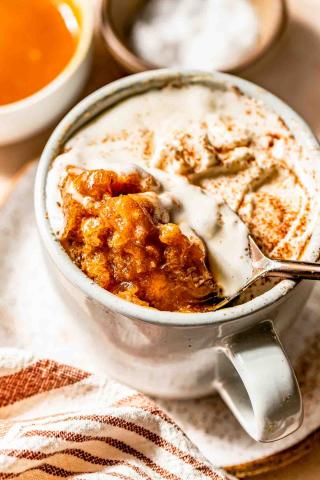 image of a standard size mug with the completed mug cake and a spoon scooping what will be a bite sized portion from the finished desert along with additonal wiped cream and ground cinnamon topping