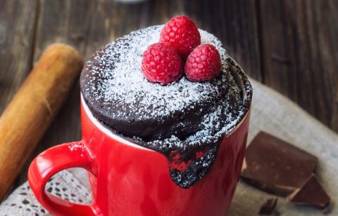 Red mug with raspberry and powdered sugar garnish on top of a chocolatey cake that is slightly above the rim of the cup to make for a delectible dessert