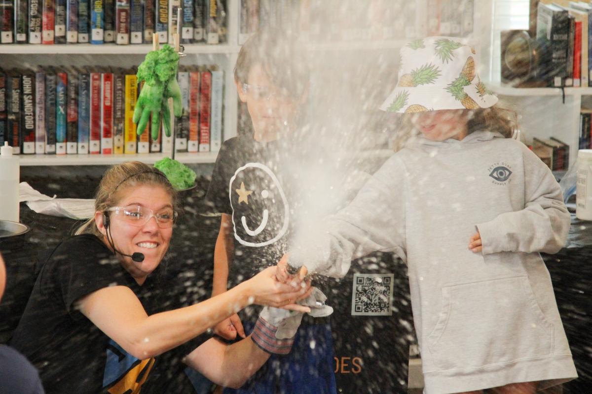 White woman with microphone pointing a cannon with two white children assisting in a library.