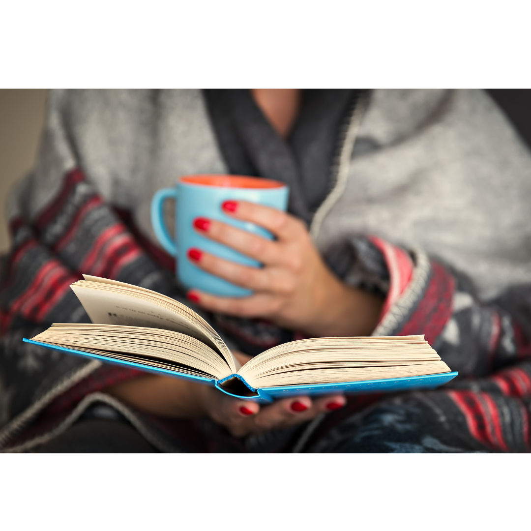 torso of a woman bundled in a blanket holding a mug in one hand and a book in the other